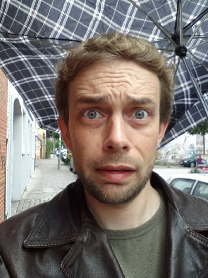 Gero Wenderholm schaut mit aufgerissenen Augen unter einem karierten Regenschirm in die Kamera, im Hintergrund eine Straße in Hamburg