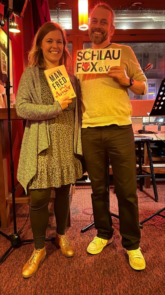 Gero Wenderholm mit Schlaufox-e.V.-Schild und einer Gästin mit dem Buch Manfred mi Amor nach der Lesung im Nachthafen